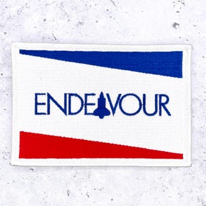 May include: A rectangular embroidered patch featuring a white background with a blue horizontal stripe at the top and a red horizontal stripe at the bottom. The text ENDEAVOUR is stitched in blue, with a blue space shuttle silhouette replacing the letter A.