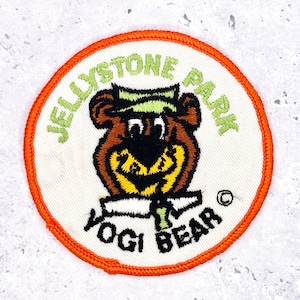 May include: Round patch featuring Yogi Bear from Jellystone Park. The patch has a white background with an orange border. The text "JELLYSTONE PARK" and "YOGI BEAR" are in green and black, respectively. Yogi Bear is brown with a yellow collar.