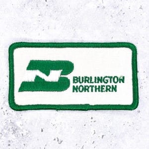 May include: A rectangular embroidered patch with a white background and a green border. It features a green stylized logo with a white letter N inside, followed by the green text BURLINGTON NORTHERN in a sans-serif font on the right side.