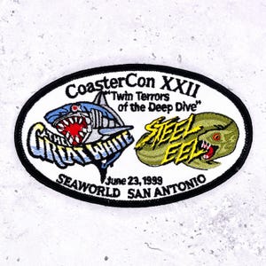 May include: An oval patch featuring a shark and an eel with open mouths. The patch reads "CoasterCon XXII" and "Twin Terrors of the Deep Dive." Below the images, it says "June 23, 1999" and "SeaWorld San Antonio."