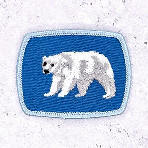 May include: Embroidered patch featuring a polar bear design. The bear is white and gray, set against a blue background with a light blue border. The patch is rectangular with rounded corners.