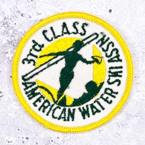 Könnte beinhalten: Runder Aufnäher mit gelbem Rand und weißem Hintergrund. Der Aufnäher zeigt eine grüne Silhouette eines Wasserskifahrers und den Text "3rd Class American Water Ski Assn."