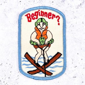 May include: A rectangular embroidered patch with a light blue border featuring a cartoon figure wearing a green helmet and orange life vest, holding a green inner tube while water skiing on brown skis over blue water, with the red text Beginner? above.