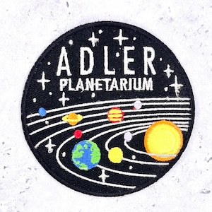 May include: A circular embroidered patch with a black background featuring the white text ADLER PLANETARIUM at the top. The design includes white orbital lines, a large yellow sun, and various colorful planets, stars, and small celestial bodies.