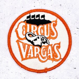 Könnte beinhalten: Runder Aufnäher mit orangefarbenem Rand und den Worten "CIRCUS VARGAS" in orangefarbener Schrift. Der Aufnäher hat einen weißen Hintergrund mit einer schwarz-weißen Illustration eines Elefanten. Über dem Text befinden sich schwarze Flaggen.