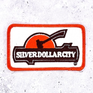 Puede incluir: Parche rectangular con borde naranja y fondo blanco. El parche presenta un diseño marrón con el texto "SILVER DOLLAR CITY" en blanco. Una forma de sol naranja está detrás del diseño.