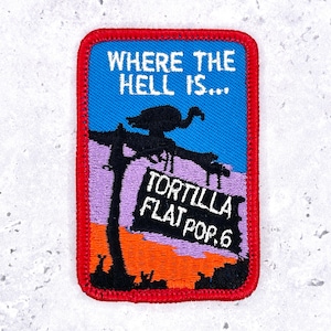 May include: A rectangular embroidered patch with a red border. The design features a blue sky, a purple mountain, and an orange landscape. It depicts a black silhouette of a bird on a branch and a sign that reads WHERE THE HELL IS... TORTILLA FLAT POP. 6.