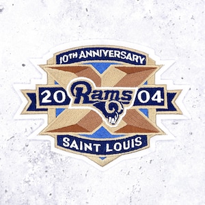 Può includere: Toppa ricamata con il logo dei St. Louis Rams, la scritta "10th Anniversary" e "2004". La toppa è blu, beige e bianca. Le parole "Saint Louis" sono anch'esse visibili sulla toppa.