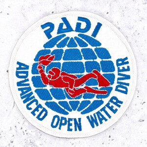 Puede incluir: Un parche bordado circular con fondo blanco y un gráfico de globo terráqueo azul. Una figura de buceador roja está centrada sobre el globo. El texto PADI aparece en la parte superior y ADVANCED OPEN WATER DIVER en azul en el borde inferior.