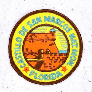 Könnte beinhalten: Runder, gestickter Aufnäher mit dem Castillo de San Marcos National Monument in Florida. Der Aufnäher hat einen gelben Hintergrund und einen braunen Rand. Blauer Text lautet "Castillo de San Marcos Nat Mon Florida". Das Fort ist orange dargestellt.