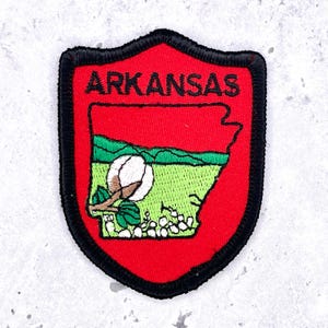 May include: Embroidered patch featuring the state of Arkansas. The patch has a red shield shape with a black border. The word "ARKANSAS" is embroidered in black. Inside the shield is a green field with a cotton plant and the state's outline.