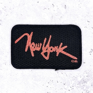 May include: A rectangular black patch with the words "New York" in red glitter script. The patch has a black border and a small "©m" marking in the lower right corner. The patch is likely for clothing or accessories.