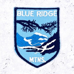 May include: A shield-shaped embroidered patch featuring a light blue background with a dark blue border. The text BLUE RIDGE is stitched in white at the top, and MTNS. is at the bottom. A dark blue pine branch and mountain silhouette are centered.