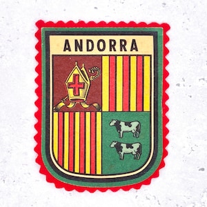 May include: A rectangular patch with a scalloped red border featuring the text ANDORRA at the top. The shield design is divided into four quadrants displaying a gold mitre with a red cross, vertical red and yellow stripes, and two black and white cows.