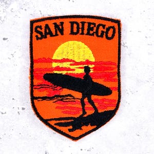 May include: A shield-shaped embroidered patch featuring the text SAN DIEGO in black letters at the top. The design depicts a black silhouette of a person carrying a surfboard on a beach against a bright yellow sun and red-orange sunset sky.