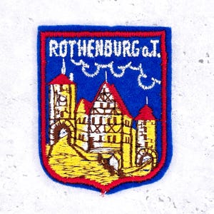 May include: Embroidered patch with a blue background and a red border. The patch features a yellow and brown illustration of a town with the text "ROTHENBURG o.T." in white.