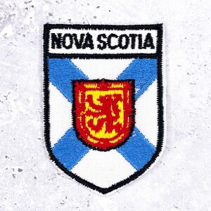 May include: Embroidered patch of the Nova Scotia flag. The patch has a white background with a blue St. Andrew's cross, a red lion rampant in the centre, and the words "NOVA SCOTIA" at the top in white.
