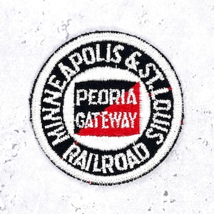 May include: A circular embroidered patch featuring a black border with white text reading Minneapolis & St. Louis Railroad. The center contains a white circle with a black and red square graphic and the white text Peoria Gateway.