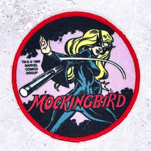 May include: Circular patch with a red trim, showcasing a comic book image of Mockingbird. The character is in a black and blue outfit, with blonde hair, wielding a weapon. The word "Mockingbird" is in red, and the Marvel Comics Group copyright is present.