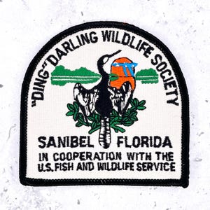 May include: A white embroidered patch with a black border featuring a black bird, a green landscape, and an orange sun. Text reads: DING DARLING WILDLIFE SOCIETY, SANIBEL FLORIDA, IN COOPERATION WITH THE U.S. FISH AND WILDLIFE SERVICE.