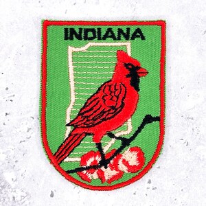 May include: A shield-shaped embroidered patch with a green background and a red border. It features a red cardinal bird perched on a black branch with white flowers. Behind the bird is a white outline of the state of Indiana with the text INDIANA.