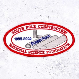 May include: Embroidered patch with a white background and a red oval border. The patch reads "SOUTH POLE CONSTRUCTION" and "NATIONAL SCIENCE FOUNDATION" in red. The center features a blue "Power Plant" graphic and the years 1999-2000.