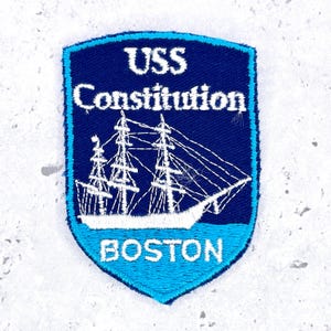May include: A shield-shaped embroidered patch featuring a dark blue background with a white sailing ship graphic. The text USS Constitution is stitched in white at the top, and the word BOSTON is stitched in white at the bottom on a light blue base.