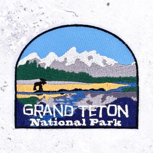 May include: A semi-circular embroidered patch featuring a landscape of Grand Teton National Park with snow-capped mountains, a green forest, a yellow meadow, and a blue lake. A black silhouette of a moose stands in the foreground with white text.