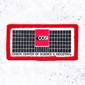 May include: Rectangular patch with a red border. The patch features a white background with a black grid pattern. A red square in the centre displays the word "COSI" in white. The text "OHIO'S CENTER OF SCIENCE & INDUSTRY" is below.