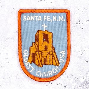 Op de afbeelding: Geborduurde patch met de tekst "SANTA FE, N.M." en "OLDEST CHURCH USA" rond een kerkillustratie. De patch heeft een lichtblauwe achtergrond en een oranje rand.