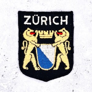 Può includere: Una toppa ricamata nera con la parola ZÜRICH in lettere maiuscole bianche in alto. Sotto il testo, due leoni gialli con lingue rosse fiancheggiano uno scudo diviso diagonalmente in bianco e blu, sormontato da una corona dorata.