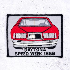 Pode incluir: Patch bordado com um carro vermelho com para-choques e faróis prateados. O texto "DAYTONA SPEED WEEK 1988" está abaixo do carro sobre um fundo cinza, emoldurado por uma borda preta.