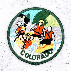 May include: A circular embroidered patch featuring three individuals in a yellow raft navigating white water rapids. They wear orange life vests and helmets. The background shows green trees and mountains. The word COLORADO is stitched in green.