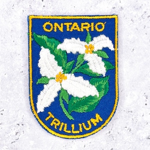 May include: Embroidered patch featuring a blue background with the words "ONTARIO" and "TRILLIUM" in yellow. The patch showcases a white trillium flower with yellow centers and green leaves. The patch has a yellow border.