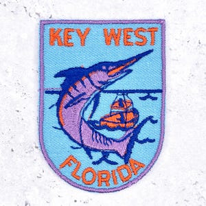 Op de afbeelding: Een schildvormige geborduurde patch met een lichtblauwe achtergrond. Het toont een paarse marlijn die boven een oranje boot springt, met de oranje tekst KEY WEST bovenaan en FLORIDA onderaan, alles omrand door een paarse gestikte rand.