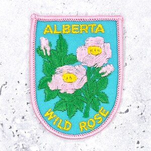 Könnte beinhalten: Ein schildförmiger, bestickter Aufnäher mit hellblauem Hintergrund und rosa gesticktem Rand. Er zeigt grüne Blätter und zwei hellrosa Wildrosen mit gelben Mitten. Der Text ALBERTA ist oben und WILD ROSE unten in Gelb aufgestickt.