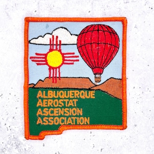 May include: Embroidered patch featuring a red hot air balloon, a yellow sun symbol, and the text "ALBUQUERQUE AEROSTAT ASCENSION ASSOCIATION." The patch has an orange border and a green lower section. The background is light blue.