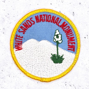 Op de afbeelding: Geborduurde ronde patch met de tekst "WHITE SANDS NATIONAL MONUMENT" in rood op een lichtblauwe achtergrond. De patch heeft een gele rand en toont een witte bergketen en een groene plant met witte bloemen.