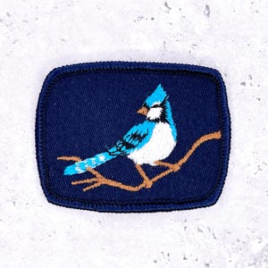 May include: Embroidered patch featuring a blue jay perched on a brown branch against a navy blue background. The bird has white, black, and turquoise details. The patch has a navy blue border and a rectangular shape with rounded corners.