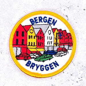 Peut inclure: Écusson brodé représentant le quai de Bryggen à Bergen, en Norvège. Le motif comprend des bâtiments rouges, jaunes et blancs, des voitures et le texte "BERGEN BRYGGEN" en bleu. L'écusson a une bordure jaune.