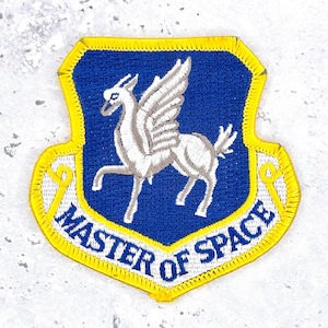 May include: A shield-shaped embroidered patch with a blue center and a bright yellow border. The patch features a white winged horse in the middle and the text MASTER OF SPACE in blue capital letters along the bottom white curved section.