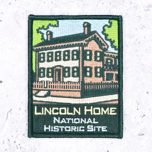 Könnte beinhalten: Ein rechteckiger gestickter Aufnäher mit einem beigefarbenen zweistöckigen Haus mit dunkelgrünem Dach und Zaun. Der Himmel ist hellblau mit grünen Bäumen. Der Text lautet LINCOLN HOME NATIONAL HISTORIC SITE in weißer und gelber Schrift auf dunkelgrünem Grund.