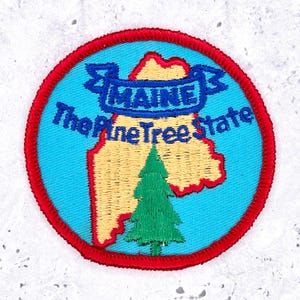 May include: A circular embroidered patch featuring a light blue background with a tan map of Maine. A green pine tree is centered at the bottom. A blue banner across the top reads MAINE, and the text The Pine Tree State is embroidered below.