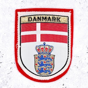 May include: A shield-shaped patch with the word "DANMARK" at the top. The patch features the Danish flag and coat of arms. The border is red with white and black stitching.