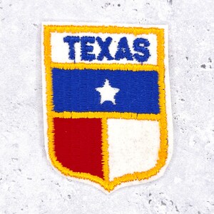 Pode incluir: Patch bordado com a palavra "TEXAS" em azul sobre um campo azul com uma estrela branca. O patch também inclui secções vermelhas e brancas, todas contornadas a ouro.
