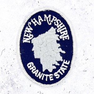 May include: An oval-shaped embroidered patch featuring a dark blue background with a white border. In the center, a white outline of the state of New Hampshire is displayed. The text NEW HAMPSHIRE is arched above, and GRANITE STATE is below.