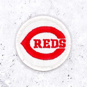 Peut inclure: Un écusson brodé circulaire avec un fond blanc et une bordure rouge cousue. Au centre, une grande lettre C rouge entoure le texte brodé rouge REDS, écrit dans une police grasse et carrée qui correspond à la couleur de la lettre C.