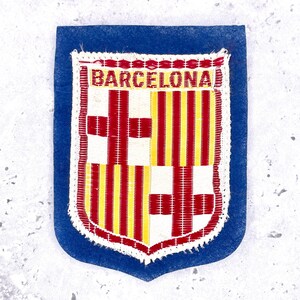 May include: A rectangular blue felt patch featuring an embroidered shield emblem with the text BARCELONA at the top. The shield displays a red cross on a white background and vertical red and yellow stripes, all bordered by white stitching.