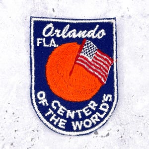Puede incluir: Parche bordado con borde azul, con el texto "Orlando FLA." y "OF CENTER THE WORLD'S". Un sol naranja y una bandera estadounidense están en el centro.