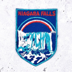 Pode incluir: Patch bordado com as Cataratas do Niágara. O patch em forma de escudo tem um fundo azul com um desenho de cachoeira branco e turquesa. O texto "NIAGARA FALLS" está em vermelho no topo, acima de um arco-íris.